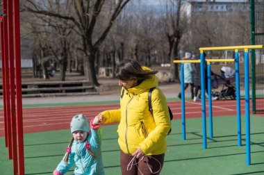 Anne, kızıyla el ele tutuşarak yürüyor. Uzun at kuyruklu bir kız. Sarı ceketli, kahverengi saçlı bir kadın. Anne ve kızı bir spor sahasında geziniyorlar..