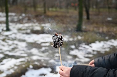 Yetişkin bir kadının eli, kış parkının önünde yanan bir kağıt çiçek tutuyor. Siyah yanmış kağıt sigara içiyor..
