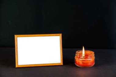Yanan bir mumun yanındaki siyah bir yüzeye karşı bej boş bir fotoğraf çerçevesi. Cam bir kavanozda kırmızı ışık. İnce çerçeveli dikdörtgen fotoğraf çerçevesinin yatay konumu.
