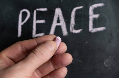 Pembe tebeşiri tutan bir adamın yakın çekimi. Barış sözcüğü arka planda siyah bir yüzeye yazılmıştır. El yazısıyla yazılmış bir metin. Önplan.