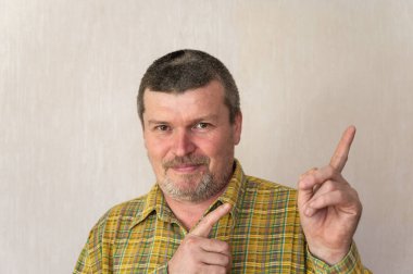 Bej bir arka planda, ekose gömlekli neşeli yetişkin bir adamın portresi. Kısa saçlı ve kır saçlı bir adam işaret parmaklarını yukarı ve yana doğru gösteriyor. Ön manzara. Kapalı.