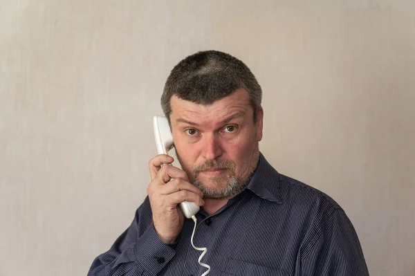 電話で話している大人の男の肖像画 青いストライプのシャツを着た男がワイヤーで白い電話受信機を持っている 灰色の短い茶色の髪 カメラを見ると — ストック写真