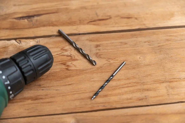 An electric drill and two drills lie against the wooden surface. The hand-held power tool is turning the boards. Selective focus. No people.