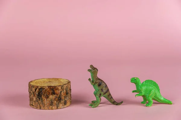 The dinosaur miniatures stand next to the pine pedestal against the pink background. Small green figures of predatory dinosaurs standing on their hind legs. Selective Focus.