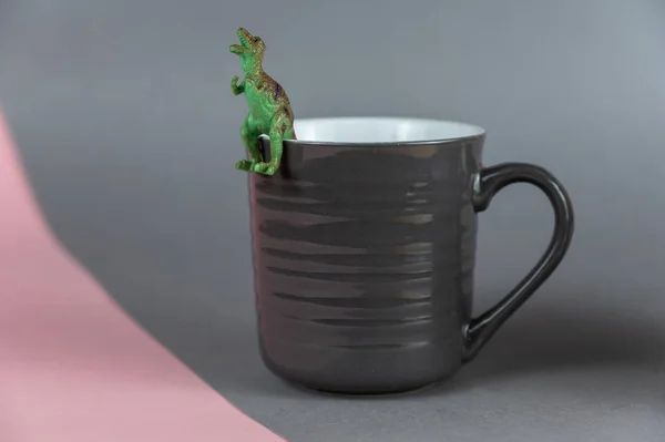 暗い灰色のマグカップの上に座っている恐竜のミニチュア 捕食恐竜の小さな緑色のフィギュア 熱い飲み物のためのハンドルを持つセラミックマグカップ グレーピンクの背景 選択的焦点 — ストック写真