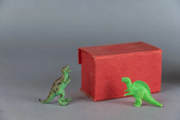 Red box and miniatures of two dinosaurs against a gray background. Small green figures of animals of prey standing on their hind legs. Rectangular closed box. Selective focus.