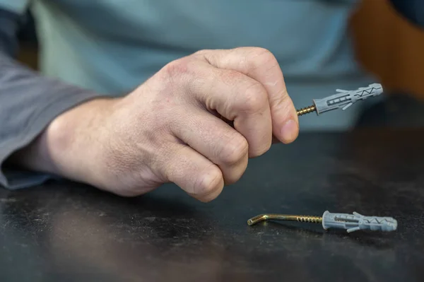 An adult male is holding an L-type screw with a dowel. A screw with a hook is used for mounting on walls, ceilings, and other surfaces. Close-up. Selective focus.