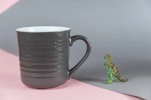 ミニチュア恐竜と灰色のピンクの背景に暗い灰色のマグカップ 前立腺恐竜の小さな緑の図は その足の上に立っている 熱い飲み物のためのハンドルを持つセラミックマグカップ 選択的焦点 — ストック写真