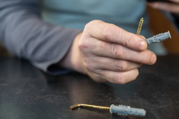 An adult male is holding an L-type screw with a dowel. A screw with a hook is used for mounting on walls, ceilings, and other surfaces. Close-up. Selective focus.