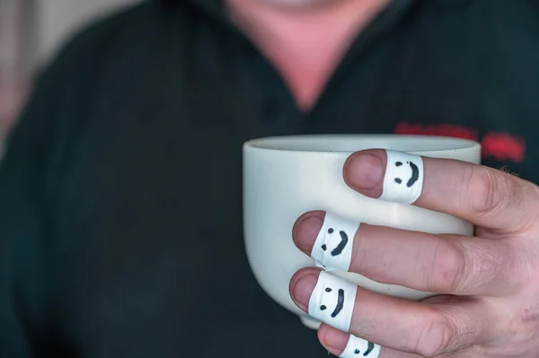 Ceramic cup in a man's hands. The first phalanges of the fingers are wrapped in a white ribbon. Smiling faces are painted on the bandages. Positive emotion concept. Selective focus. Close-up