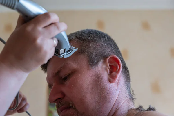 Haircut for a grown man. A woman's hand with an electric hair clipper cuts short, graying hair. A middle-aged person with a naked torso sits inside a living room. Series part. Selective Focus