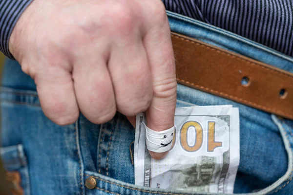 A man\'s hand pulls money out of his jeans pocket. The index finger is wrapped in white tape. A happy smiling face is drawn in black marker on the finger. One hundred American dollars.