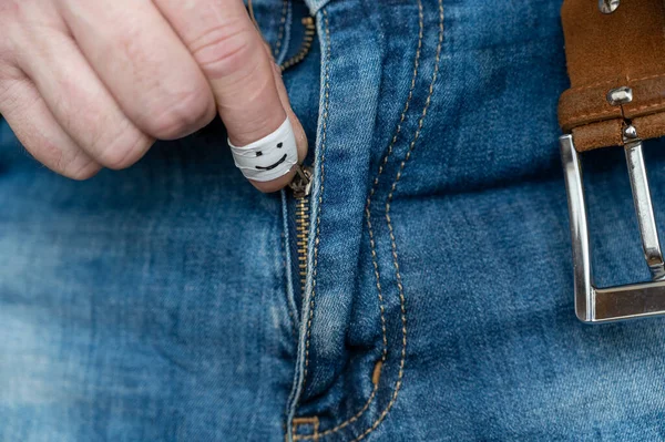 A man\'s hand unzips the fly of his blue jeans. The index finger is wrapped in white tape. A happy smiling face is drawn in black marker on the finger. Close-up. Selective focus.