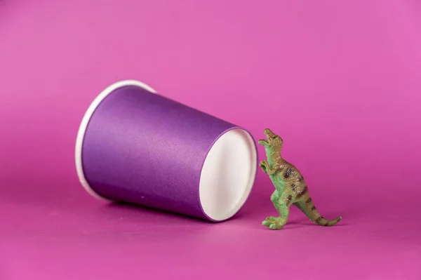 A miniature dinosaur and a paper cup against a purple background. A lilac disposable drink glass. Green figure of a predatory animal standing on its hind legs. Selective focus.