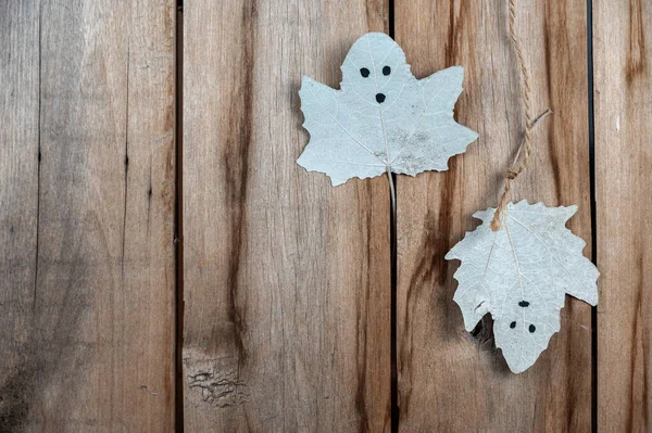 Leaf of a tree with ghost faces drawn against a wooden background. White poplar leaves with drawn eyes and mouth hanging vertically on string. Background with copy space for text and design elements