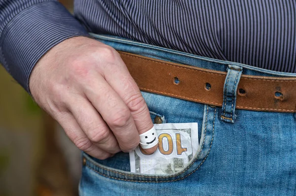 A man\'s hand pulls a hundred dollars from his pant pocket. The index finger is wrapped in white tape. A happy smiling face is drawn in black marker on the finger. Selective Focus.