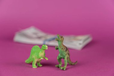 Dinozor minyatürleri ve mor arka planda bir tomar banknot. Arka planda bir tomar para var. Arka ayaklarının üzerinde duran yırtıcı hayvanların yeşil figürleri. Seçici odak.