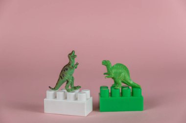 Çocuk müteahhitinin iki bloğunda duran minyatür dinozorlar. Arka ayaklarının üzerinde duran iki küçük yeşil yırtıcı dinozor minyatürü. Pembe arka plan. Seçici Odaklanma.