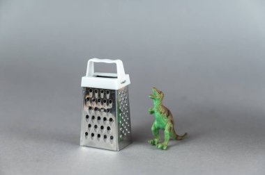 Gri arka planda küçük rende ve minyatür dinozor var. Yırtıcı bir dinozorun arka ayakları üzerinde duran küçük yeşil figürü. Beyaz saplı metal rendesi. Seçici odak.