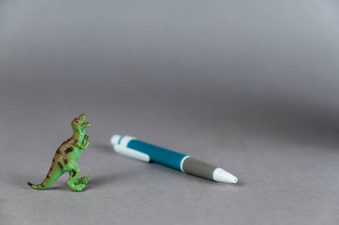 Gri arka planda bir dinozor ve tükenmez kalem minyatürü. Arka ayakları üzerinde duran yırtıcı bir hayvanın küçük yeşil biblosu. Beyaz-gri-mavi plastik kalem. Seçici odak.