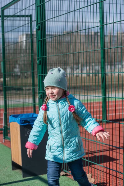 Portrait of a child having fun on a sports field. A five-year-old girl with long hair. Hair braided into pigtails. The child is dressed in warm clothes. Childhood. Outdoors. A sunny spring day.