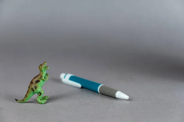 一只恐龙和一支圆珠笔在灰色背景下的缩影 食肉动物的绿色小雕像 后腿站立着 白灰蓝色塑料笔 有选择的重点 — 图库照片