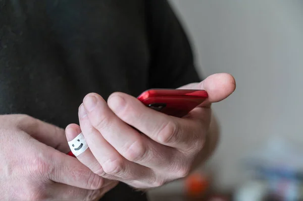 一个男人拿着一部红色的智能手机 他小指的第一个指节用白色缎带包裹着 绷带上画着一张笑脸 积极的情感概念 选择性焦点 — 图库照片
