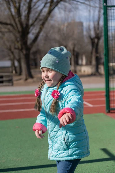 Portrait of a child having fun on a sports field. A five-year-old girl with long hair. Hair braided into pigtails. The child is dressed in warm clothes. Childhood. Outdoors. A sunny spring day.