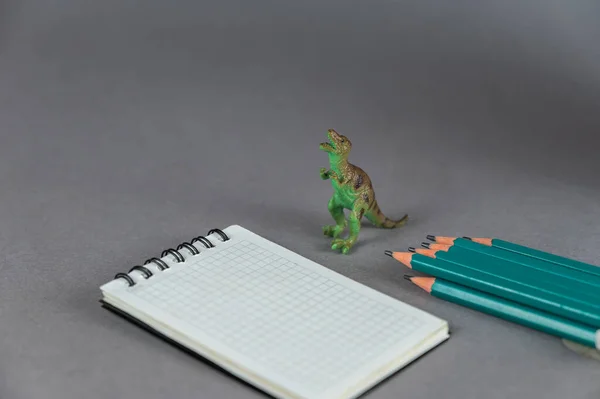 A miniature dinosaur, a set of pencils and a notebook against a gray background. Small green figure of an animal of prey standing on its hind legs. Spring-loaded caged notebook