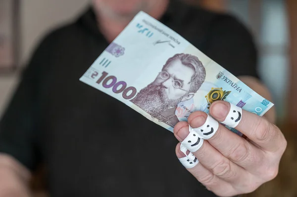 A grown man is holding money in his hand. One thousand Ukrainian hryvnias. The first phalanges of his fingers are wrapped in white ribbon. Smiling faces are painted on the bandages.