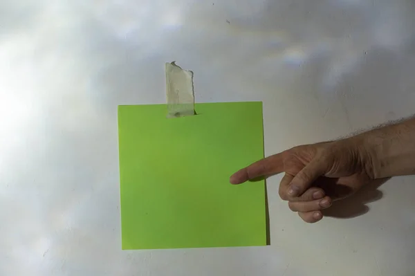 The index finger of the man\'s hand points to a blank green square sheet of paper fixed to the wall. An abstract light and shadow drawing on a gray plastered wall.  Selective focus. Low key.