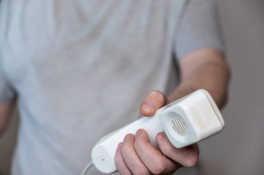 Orta yaşlı bir adam bir telefon ahizesi tutuyor. Bir adamın elinde beyaz plastik bir telefon ahizesi. Gri tişörtlü olgun bir adamın orta kısmı. Odanın içinde. Seçici Odaklanma.