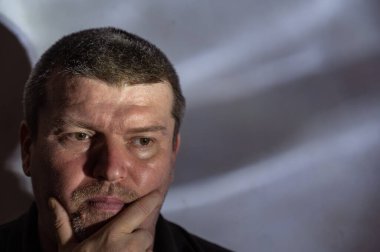 Orta yaşlı bir adamın gri duvardaki portresi. Tıraşsız bir adam çenesini elinde tutar. Kısa kır saçlı, kahverengi saçlı. Duvardaki soyut gölgeler. Kameranın arkasına bakıyorum. Seçici odaklanma. Alçak tuş