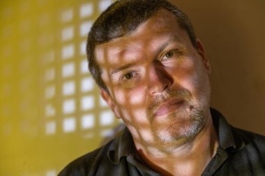 Sarı duvara karşı barışçıl bir adamın alışılmadık portresi. Tekrarlanan kare ışık desenleri. Işık ışınları yüzünü kısmen aydınlatıyor. Sakin suratlı adam kameraya bakıyor..