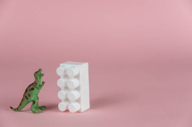 Dinozor minyatürü, pembe arka planın karşısındaki çocuk inşaatçısının beyaz bloğunun yanında duruyor. Yırtıcı bir dinozorun arka ayakları üzerinde duran küçük yeşil minyatürü..