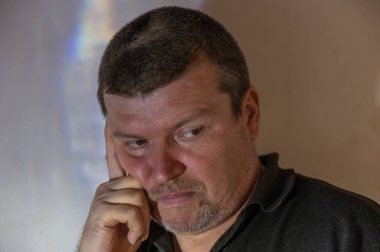 Düşünceli bir adamın portresi. Tıraşsız erkek yanağını avucuna koyar. İşaret parmağı şakağında duruyor. Orta yaşlı, kısa gri saçlı bir erkek. Kameranın ötesine bakıyorum..