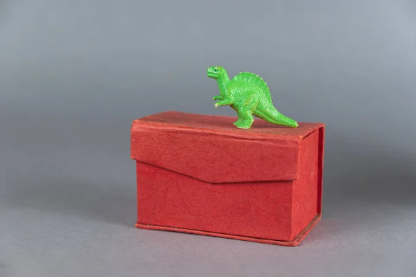 Red box and miniature of one dinosaur against a gray background. Small green figurine of an animal of prey standing on its hind legs. Rectangular closed box. Selective focus.