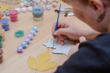 Sanatçı kurumuş yapraklarla resim çizer. Bir kadının elleri kavak yaprağının üzerine siyah guaj boyasıyla fırçalar. Masada akrilik ve guaj var. İçeride. Seçici odak.