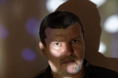 Duvara dayanmış sessiz bir adamın portresi. Bir ışık demeti sağ gözünü aydınlatıyor. Orta yaşlı, kısa gri saçlı bir erkek. Adam kendinden emin bir şekilde kameraya bakıyor. Seçici odak.