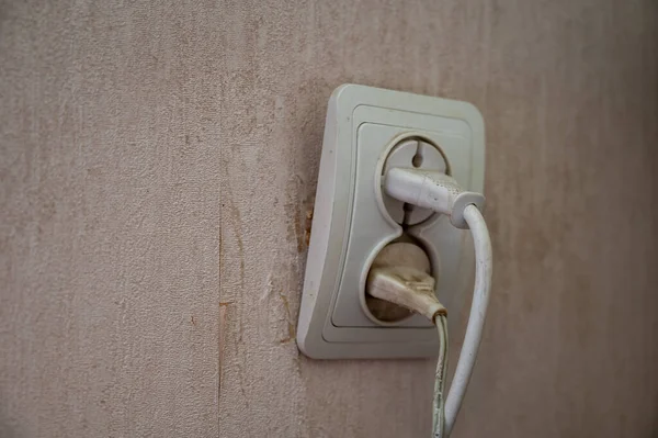Broken electrical outlet in beige wall. White electrical outlet with two plugs connected. Danger of electric shock. Possibility of fire due to a short circuit.  Selective focus.