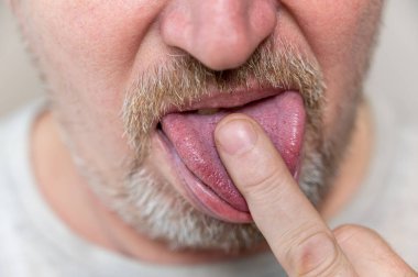 Bir erkeğin yüzünün alt kısmı. Sakallı, olgun bir adam. Orta parmağını diliyle yalıyor. Sakalında ve bıyığında gri saç var. Adam kameranın önünde kıvranıyor. Yakın plan. Seçici odak.