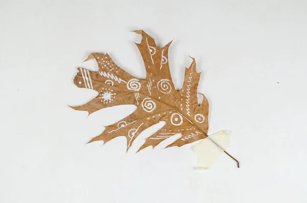 Ethnic painting on a yellow fallen leaf. White abstract patterns on dry leaf attached to the wall with duct tape. Painting with paints on fallen leaves. Contemporary art. Talent. Selective focus.