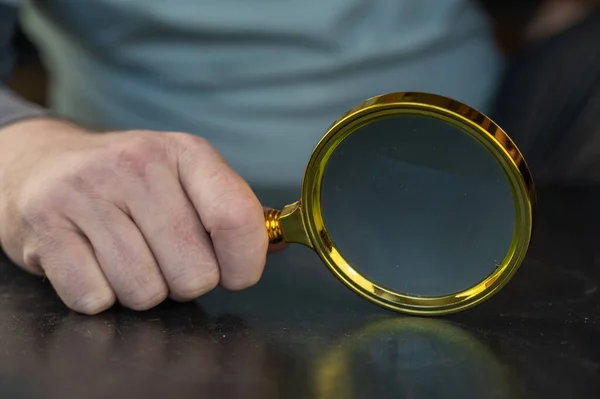 Grown man holding a magnifying glass. A large round loupe in a gold-colored frame. The man is sitting at a black table inside the room. Close-up. Selective focus.
