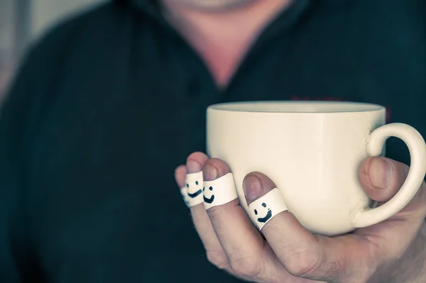 Ceramic cup in a man\'s hands. The first phalanges of the fingers are wrapped in a white ribbon. Smiling faces are painted on the bandages. Positive emotion concept. Selective focus. Close-up