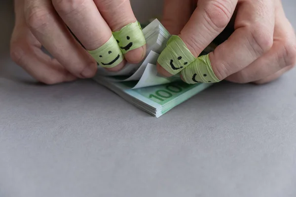 A man's fingers are counting the money. A wad of euro bills. The index and middle fingers of both hands are wrapped in green tape. Happy smiling faces are drawn on the fingers.
