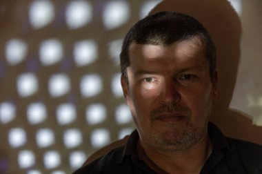 Düşünceli bir yüzü olan orta yaşlı bir adamın portresi. Adamın sırtı duvara dönük. Duvarda ve adamın yüzünde tekrarlayan şekillerin ışık ve gölge deseni. İnsanlar, duygular