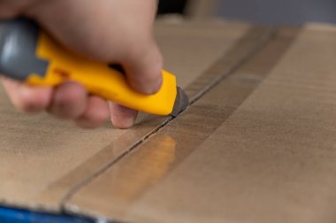 Bir adamın eli, sarı keski ile karton bir kutuyu açar. Paketin flapları yapışkan bantla kapatılmış. Yakın plan. Seçici odak.