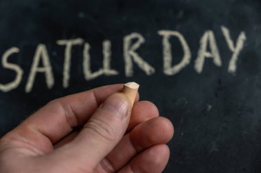 An adult man holds a piece of yellow chalk in his hand. Handwritten word SATURDAY on black chalkboard. Days of the week. Calendar