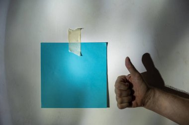 Olgun bir adamın eli iyi bir jest gösterir. Duvara sabitlenmiş boş mavi kare kağıt. Gri alçılı bir duvara soyut bir ışık ve gölge çizimi. Seçici odaklanma. Alçak tuş.