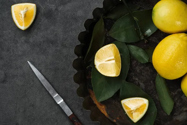 Limonlar. Taze olgun sarı turunçgil meyveleri ve metal bir kapta yapraklı limon dilimleri. Koyu renkli masada bıçaklı meyvelerin üst görüntüsü